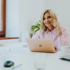 Kobieta w różowej koszuli rozmawia przez telefon przy biurku z laptopem.