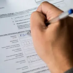 Dłoń pisze na formularzu "Zgłoszenie pobytu stałego", wpisując nazwisko. Dokument ten jest kluczowy do uzyskania tytułu prawnego do lokalu.