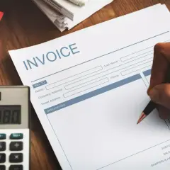 Ręka pisze na fakturze "INVOICE". Obok leży kalkulator. Faktura VAT marża - co oznacza dla kupującego?