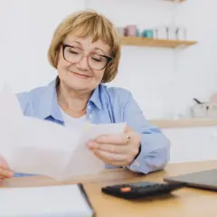 Uśmiechnięta starsza pani analizuje dokumenty, być może dotyczące tego, renta wdowia komu przysługuje. Na stole leży kalkulator i laptop.