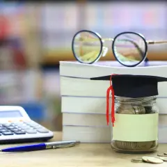 Skarbonka w kształcie biretu studenckiego, pełna monet, obok stosu książek, kalkulatora i okularów. Idealna na umowa zlecenie dla studenta.