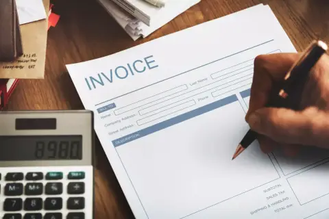 Ręka pisze na fakturze "INVOICE". Obok leży kalkulator. Faktura VAT marża - co oznacza dla kupującego?