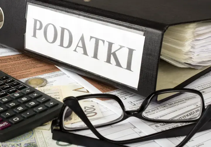 Segregator z napisem "PODATKI", okulary, kalkulator i pieniądze na dokumentach. Wszystko gotowe do rozliczenia rezydencji podatkowej.