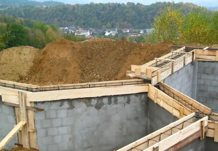 Fundamenty domu w budowie, zbrojenie i szalunki. Widać kopce ziemi i zabudowania w tle. Ważne jest pozwolenie na budowę, by wszystko szło zgodnie z planem.