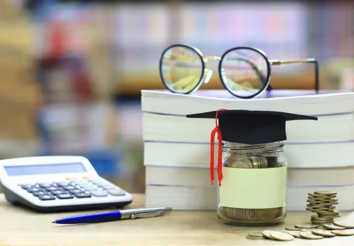 Skarbonka w kształcie biretu studenckiego, pełna monet, obok stosu książek, kalkulatora i okularów. Idealna na umowa zlecenie dla studenta.