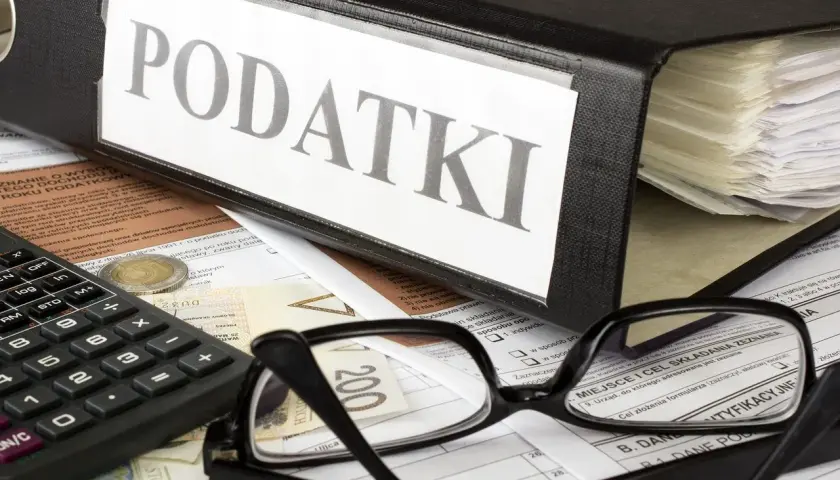 Segregator z napisem "PODATKI", okulary, kalkulator i pieniądze na dokumentach. Wszystko gotowe do rozliczenia rezydencji podatkowej.