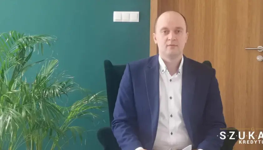 Mężczyzna w garniturze siedzi w fotelu obok rośliny, na tle zielonej ściany i drewnianych drzwi.