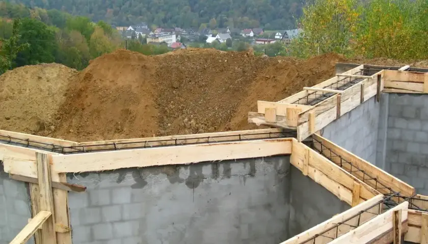 Fundamenty domu w budowie, zbrojenie i szalunki. Widać kopce ziemi i zabudowania w tle. Ważne jest pozwolenie na budowę, by wszystko szło zgodnie z planem.
