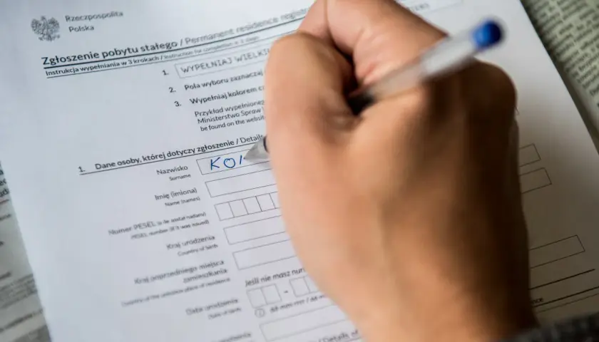 Dłoń pisze na formularzu "Zgłoszenie pobytu stałego", wpisując nazwisko. Dokument ten jest kluczowy do uzyskania tytułu prawnego do lokalu.