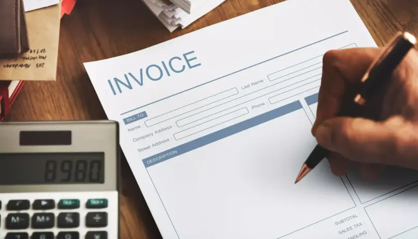 Ręka pisze na fakturze "INVOICE". Obok leży kalkulator. Faktura VAT marża - co oznacza dla kupującego?