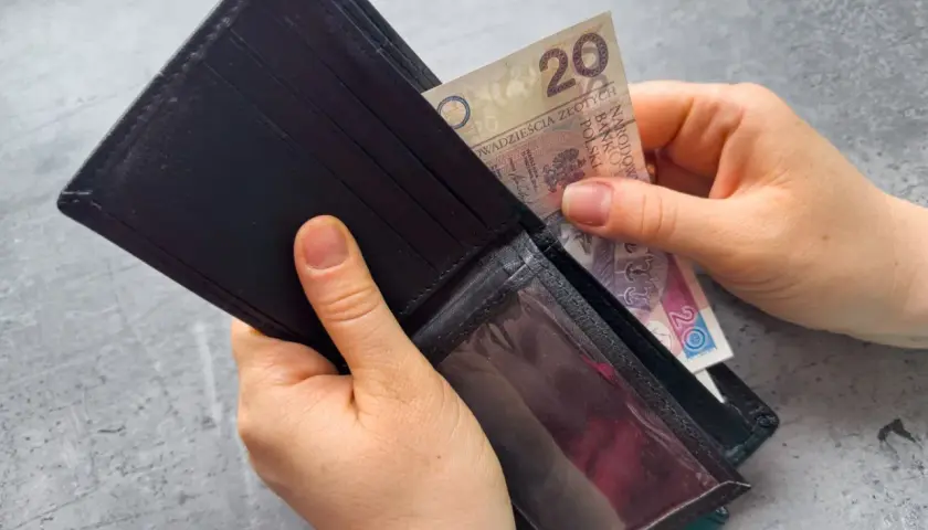 Dłonie wkładają banknot 20 zł do czarnego portfela. Zastanawiasz się, po jakim czasie od zarejestrowania wypłacany jest zasiłek dla bezrobotnych?