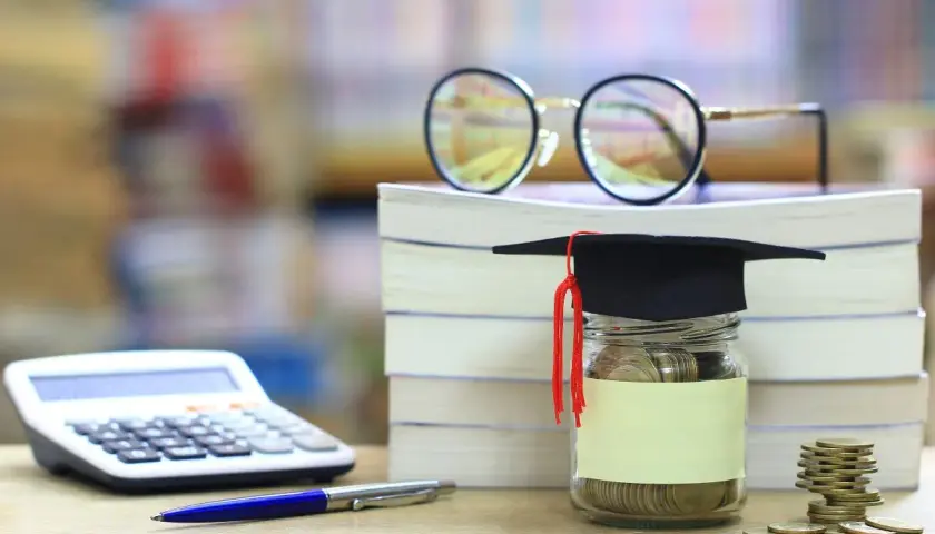Skarbonka w kształcie biretu studenckiego, pełna monet, obok stosu książek, kalkulatora i okularów. Idealna na umowa zlecenie dla studenta.