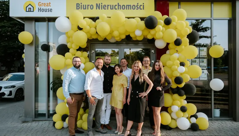 Grupa ludzi pozuje przed biurem nieruchomości ozdobionym żółtymi, czarnymi i białymi balonami.