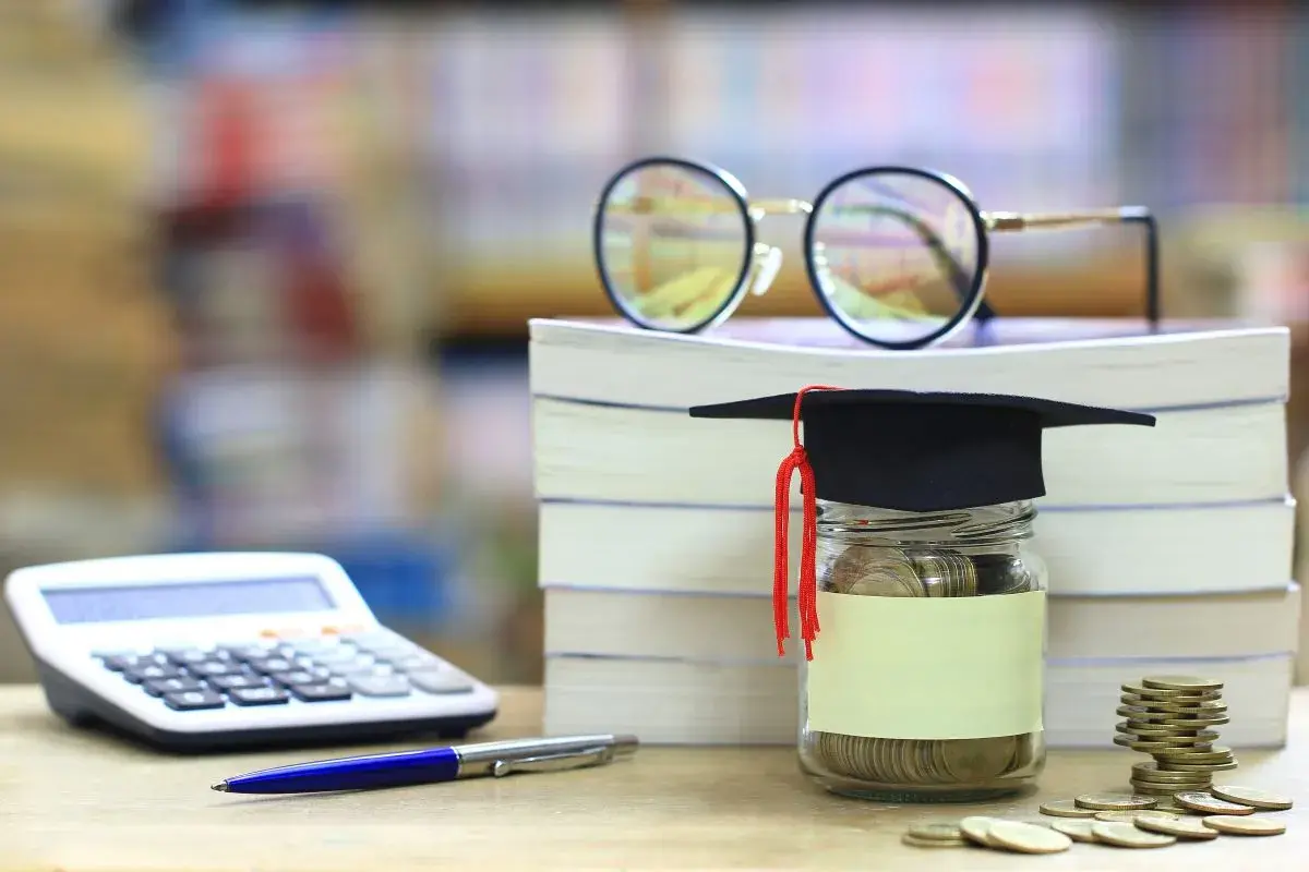 Skarbonka w kształcie biretu studenckiego, pełna monet, obok stosu książek, kalkulatora i okularów. Idealna na umowa zlecenie dla studenta.
