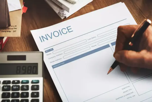 Ręka pisze na fakturze "INVOICE". Obok leży kalkulator. Faktura VAT marża - co oznacza dla kupującego?